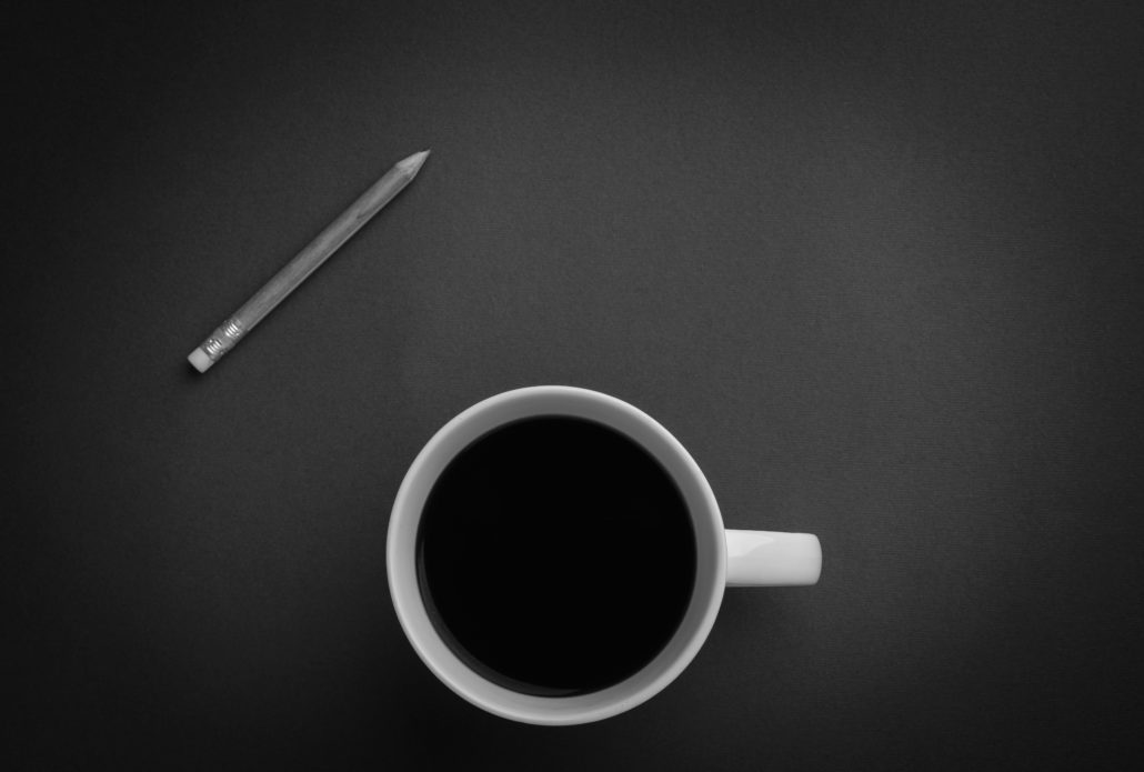 Black and white overhead photo of a pencil and a mug of black coffee on a plain dark surface, calm and simple mood.